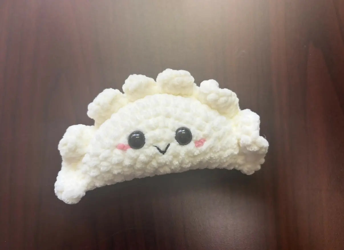 A white crocheted dumpling shaped plush toy with black button eyes and pink cheeks representing perogy, related to canadian made skincare