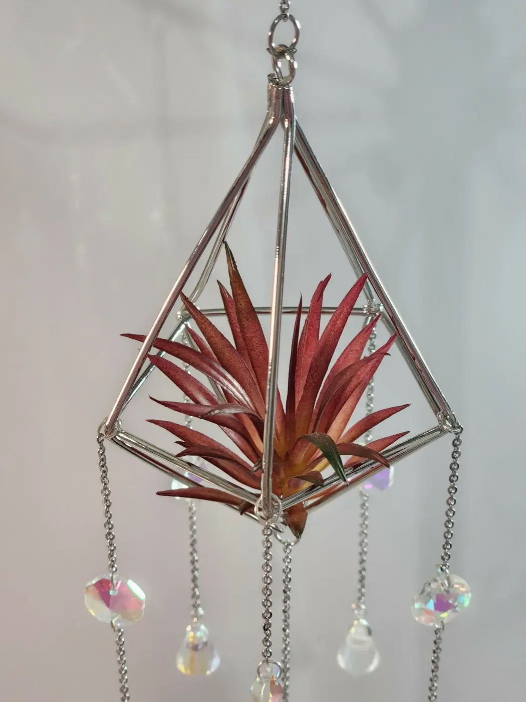 A geometric silver-toned metal pyramid air plant suncatcher with dangling crystal beads and a red green air plant