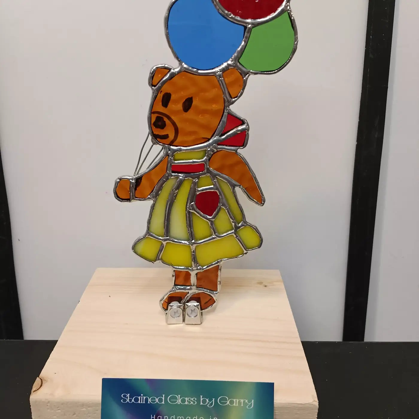 Handmade locally colorful stained glass teddy bear in yellow dress with red accents holding blue green and red bear w balloons