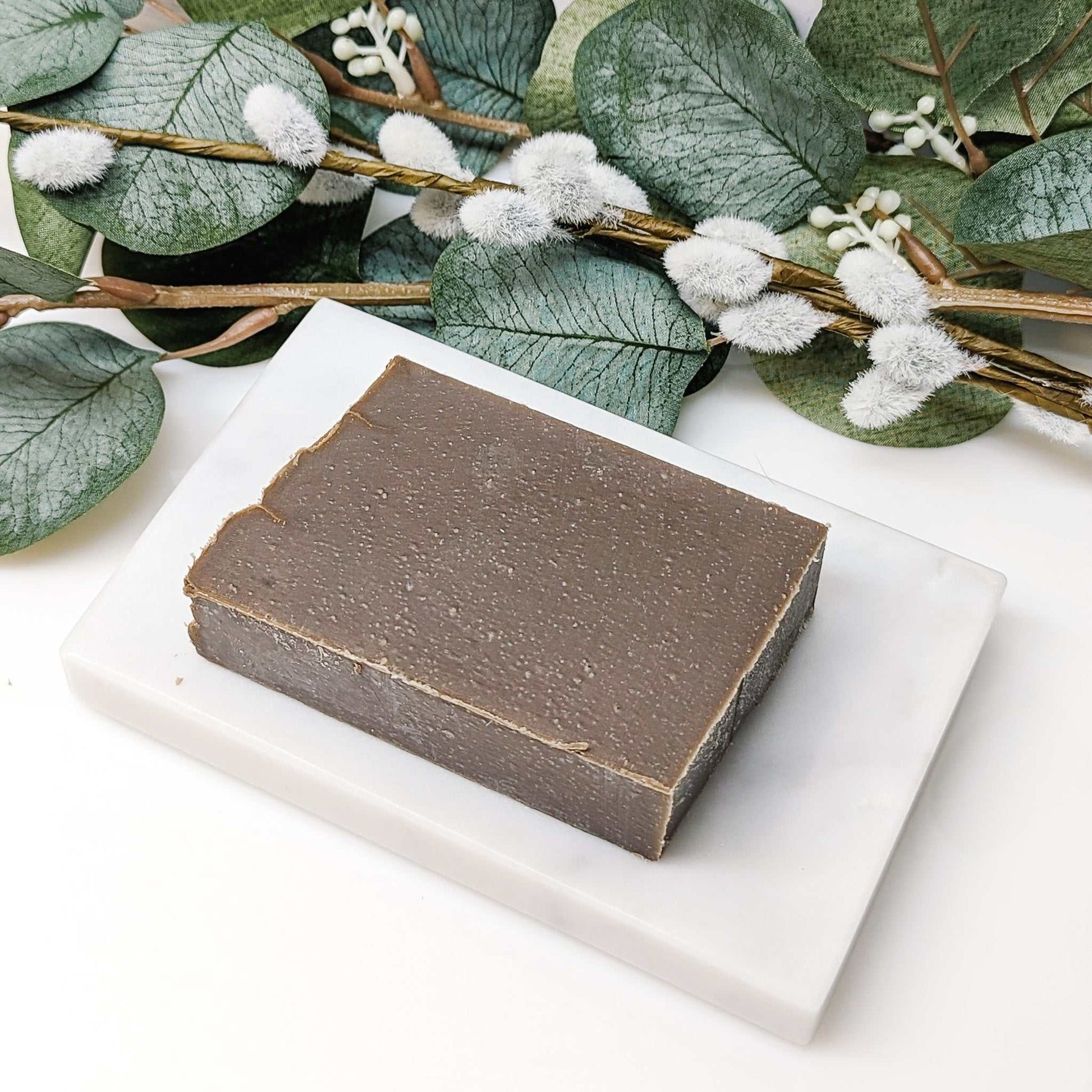 Coffee Soap Bar on a white tray surrounded by greenery, crafted from organic coffee and nourishing oils for smooth, hydrated skin.