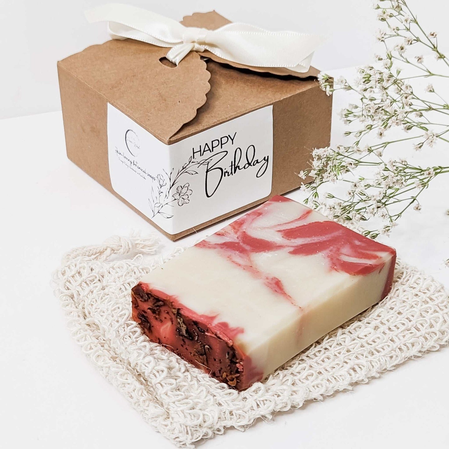 Handcrafted soap bar with swirls of red and white, paired with a rustic brown gift box featuring a 'Happy Birthday' tag, all set against a white backdrop with delicate white flowers.