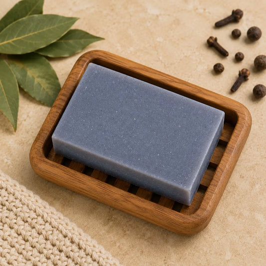 A solid blue bay rum soap bar made with sodium shea butterate rests in a wooden soap dish with slotted sides