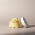 A clear glass jar with a white lid filled with creamy pale yellow whipped body butter with cocos nucifera and theobroma cacao