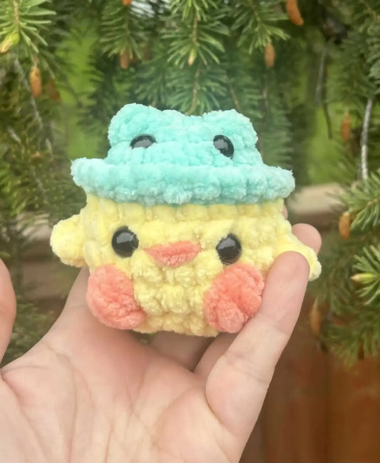 A soft crocheted yellow duck craft with pink cheeks wearing a frog-shaped hat made from pure wash yarn