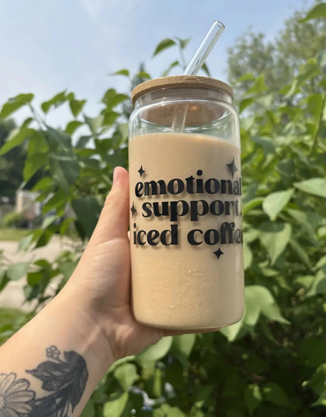 A clear glass iced coffee cup with wooden lid and emotional support iced vinyl decal filled with light brown beverage