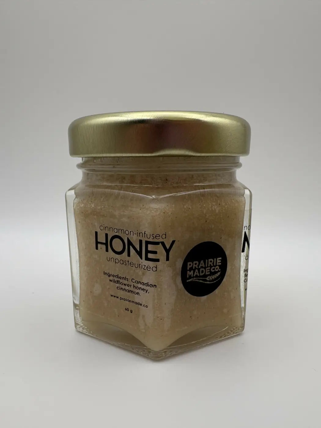 A hexagonal glass jar with a gold lid containing cinnamon-infused unpasteurized honey from prairie made co flavoured honey trio