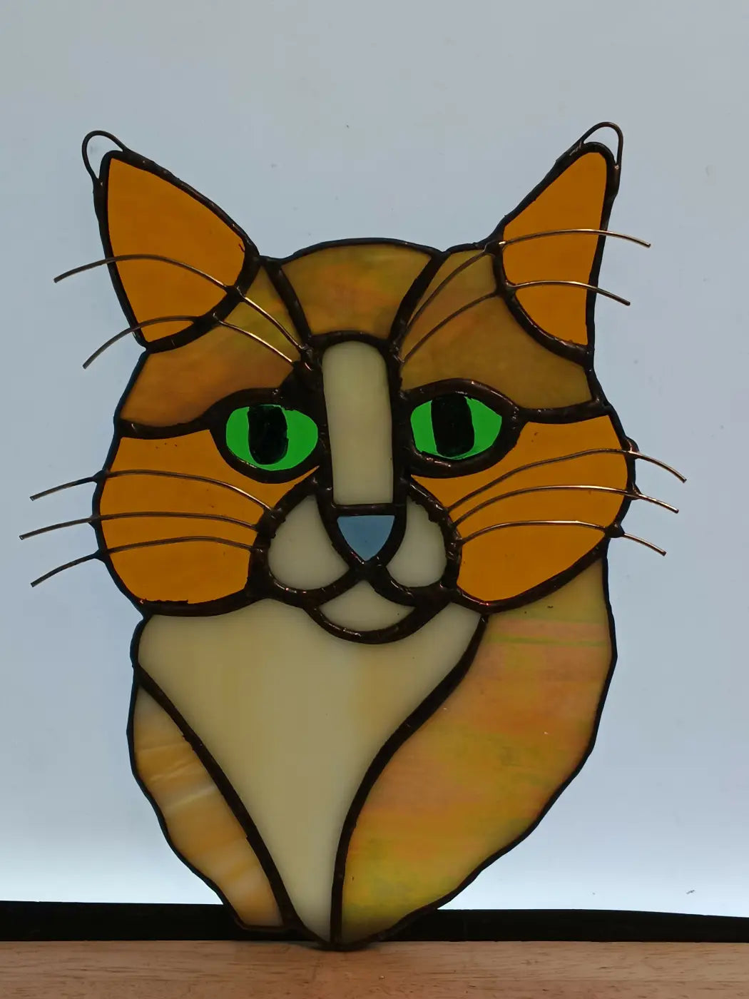 A vibrant orange and cream stained glass cat headshot with bright green eyes in a traditional leaded design