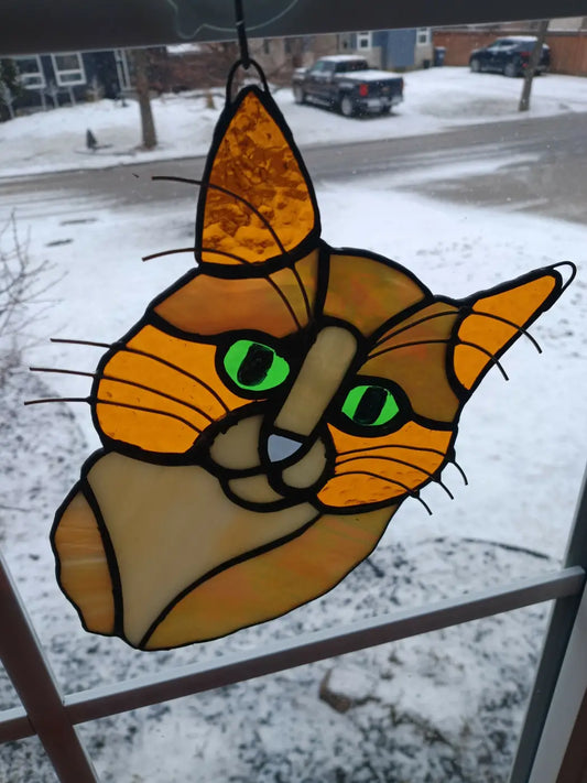 A vibrant orange and tan stained glass cat headshot ornament with striking green eyes for decorative use