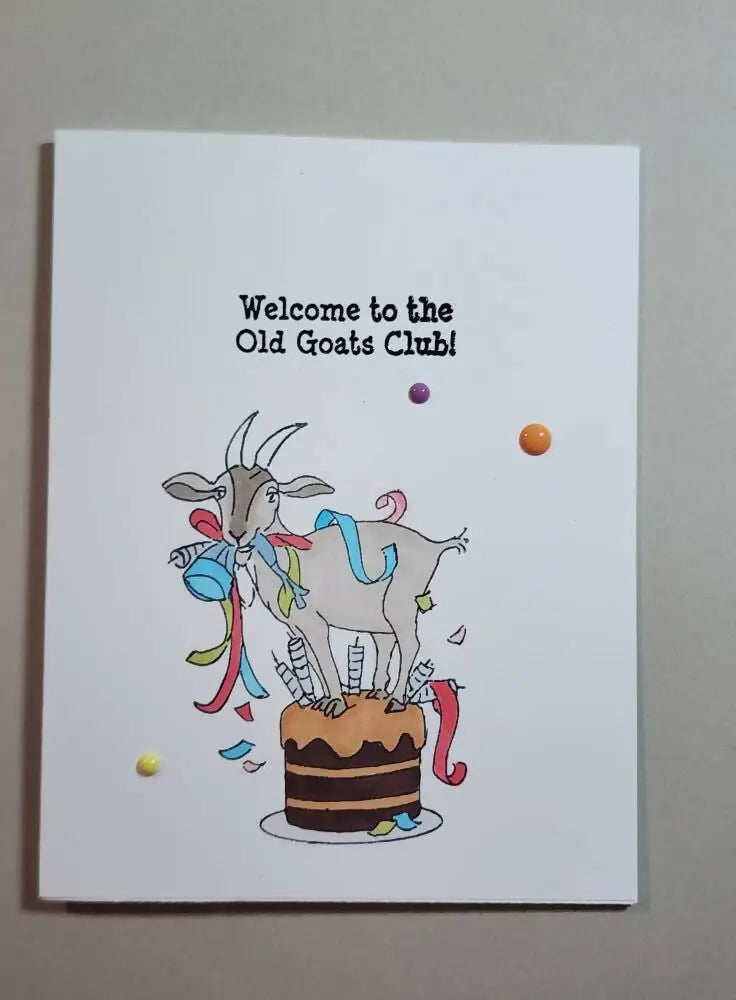 A whimsical white greeting card featuring a cartoon goat on a chocolate cake with colorful ribbons and candles