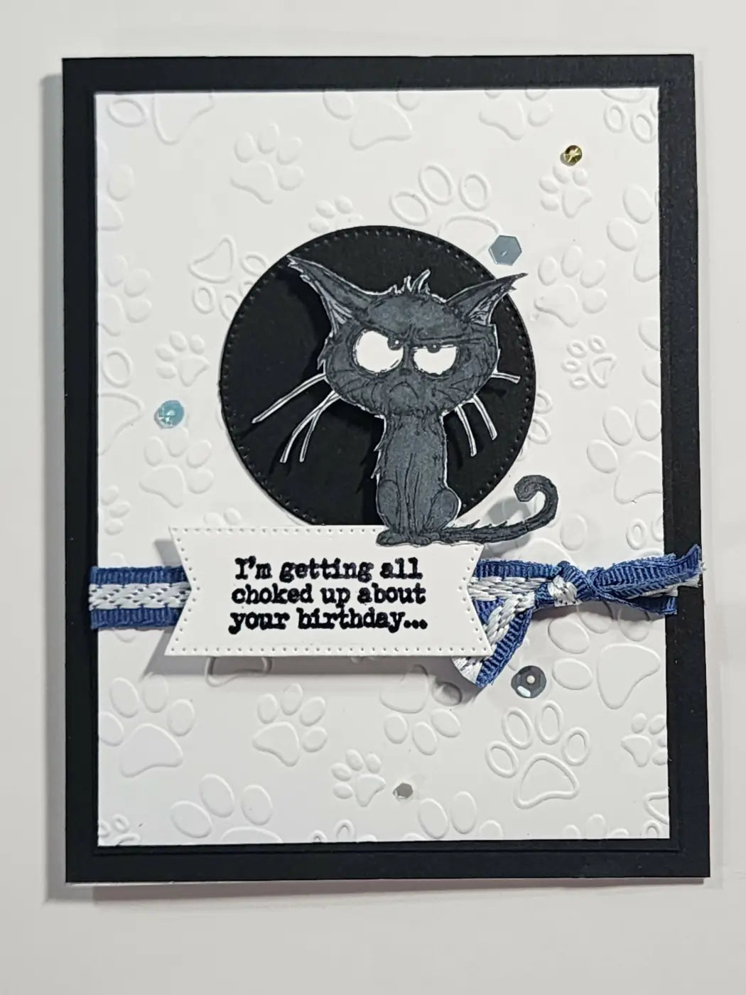 Black and gray cartoon cat with angry eyes in circular cutout on white greeting card with embossed paw prints perfect for canadian made skincare gifts