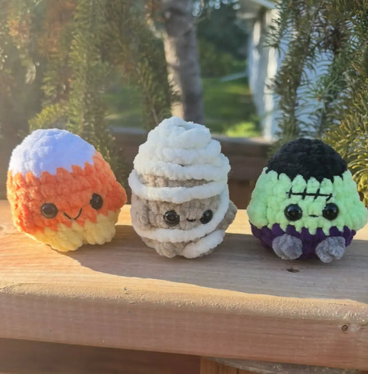 Three handmade crocheted halloween palls featuring candy corn, mummy, and frankenstein displayed on wooden surface