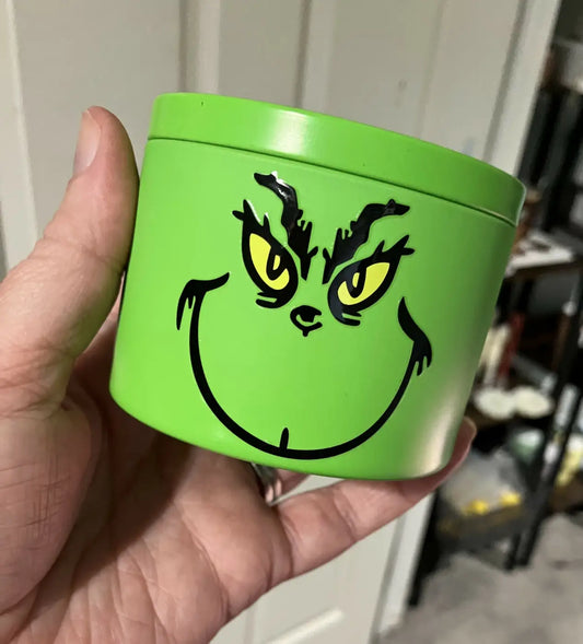 Bright green specialty character candle tin featuring a black and yellow grinch face with clean-burning holiday soy wax