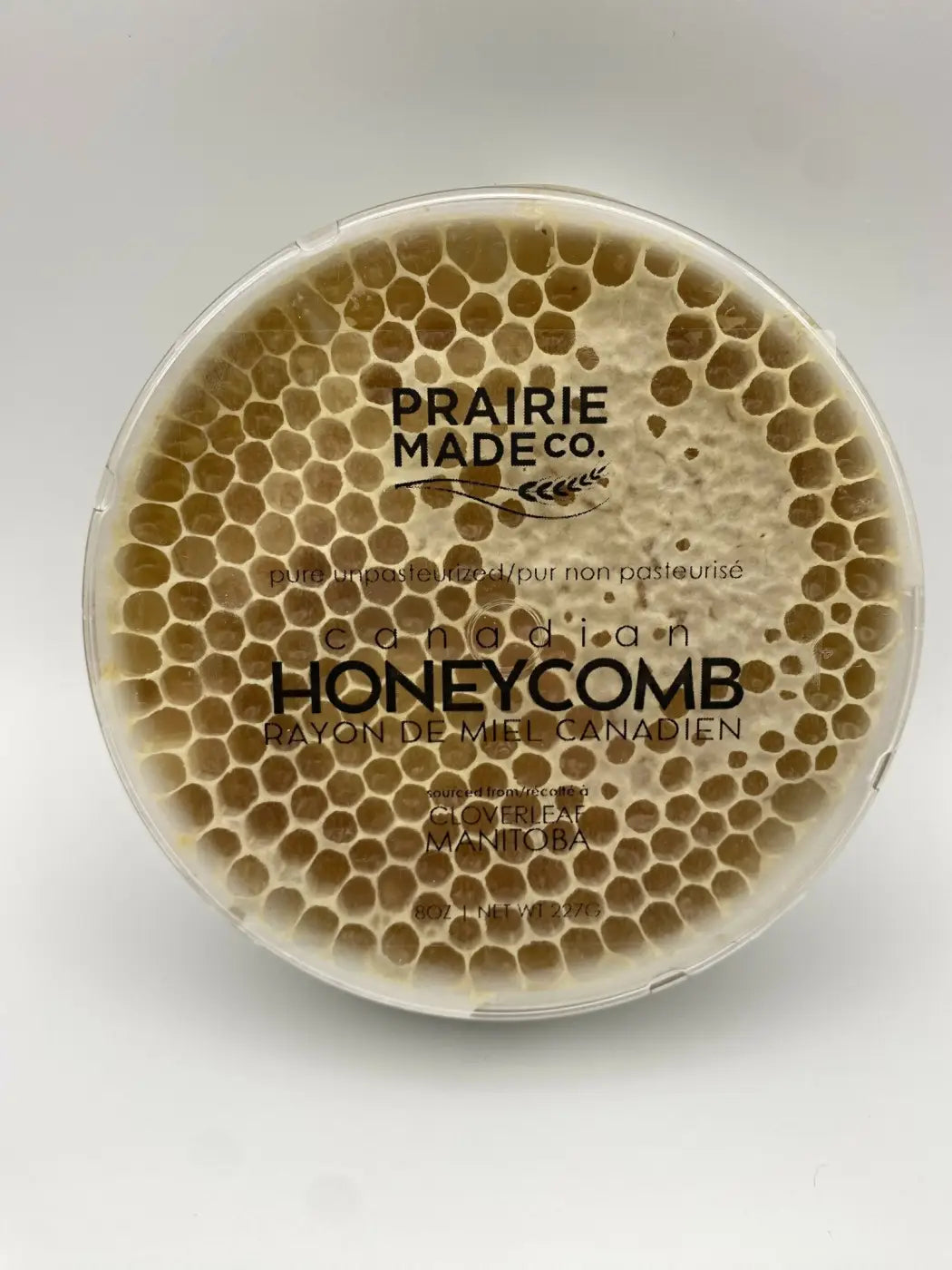 A round transparent container holds prairie made co honeycomb with golden brown hexagonal cells of locally sourced honey candy