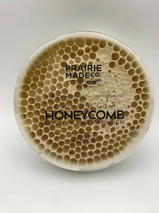 A round transparent container holds prairie made co honeycomb with golden brown hexagonal cells of locally sourced honey candy
