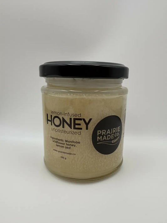 A clear glass jar with a black lid contains 250g of prairie classic lemon-infused honey with a fragrant twist