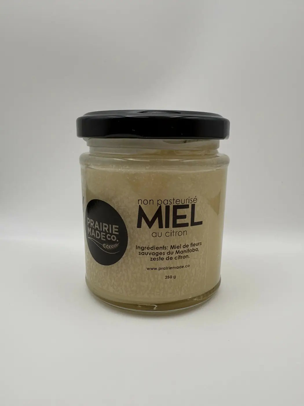 A clear glass jar with black lid contains pale yellow lemon honey labeled miel au citron by prairie nadeco with fragrant twist