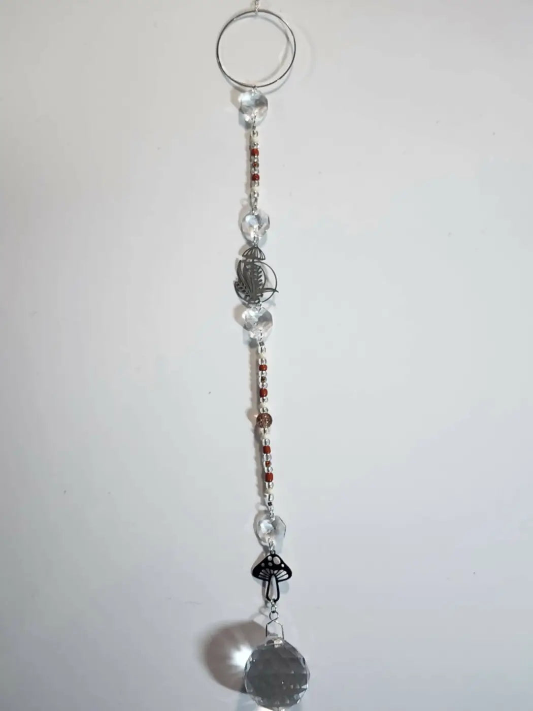 Long suncatcher with faceted crystal, silver ring, red beads, leaf charm, and black mushroom pendant for direct sunlight display