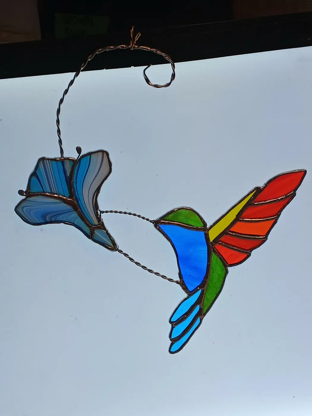 A vibrant stained glass midflight hummingbird with blue body and rainbow wings suspended by twisted wire hanger
