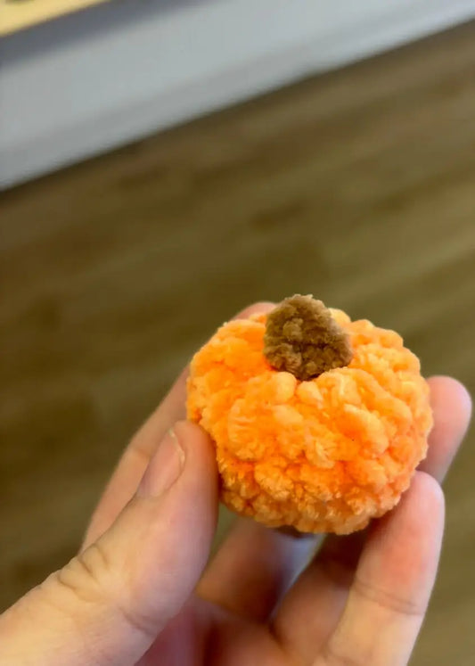 A hand holds a small orange handmade crochet mini pumpkin with a brown stem natural skincare Canada accessory