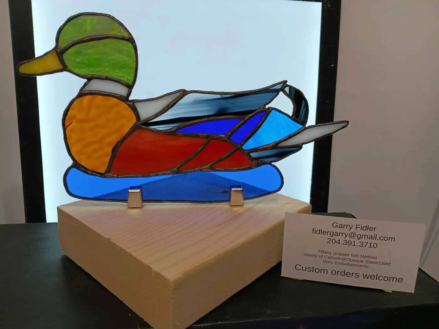 A multi colored craft stained glass duck with green head orange body blue wings on a wooden base handmade stained glass
