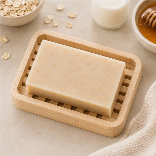 A light beige rectangular oatmeal milk and honey soap bar with speckled texture in a natural wood slatted soap dish
