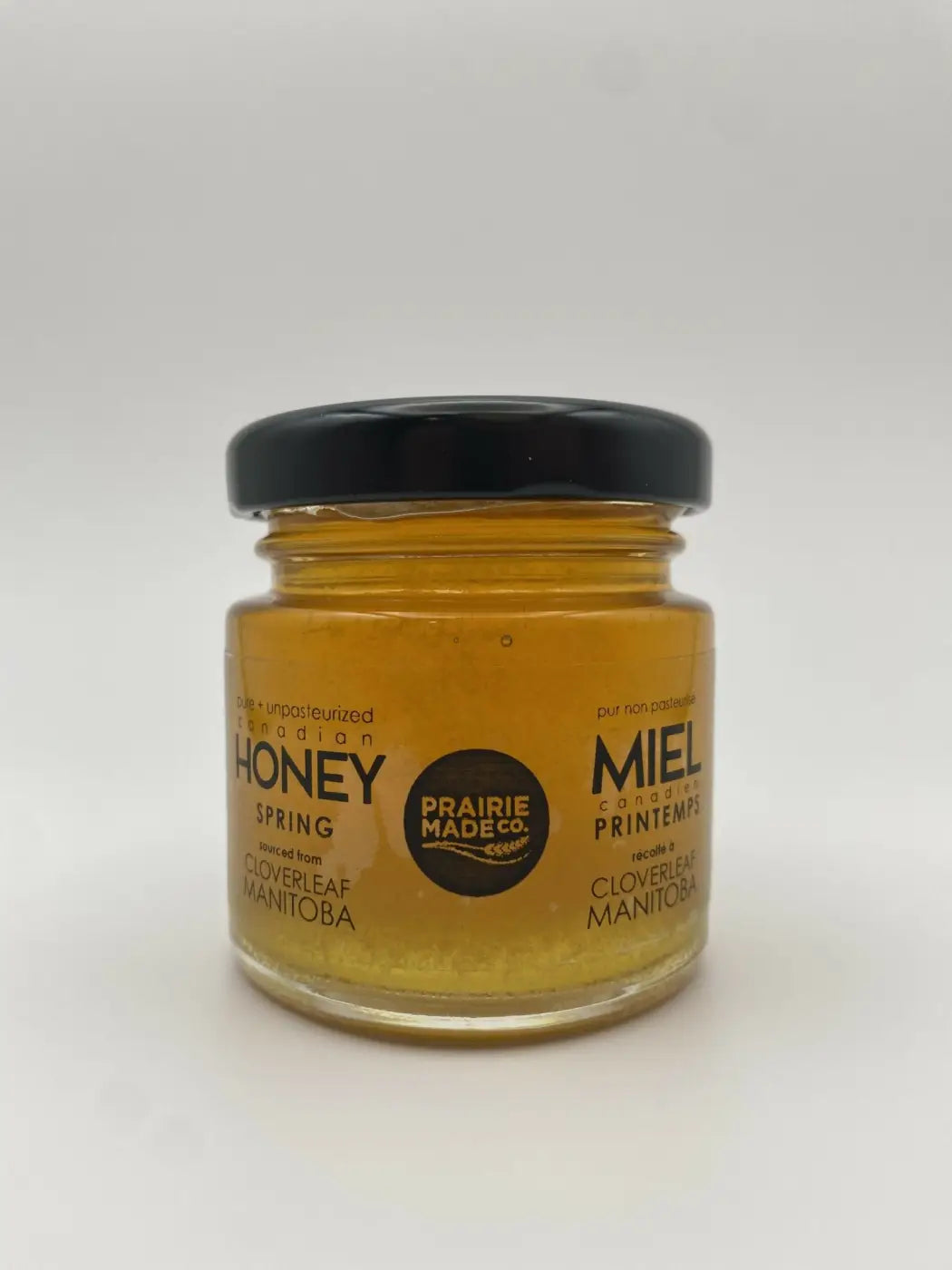 A clear glass jar with black lid contains golden honey labeled honey spring from prairie made season honey sampler