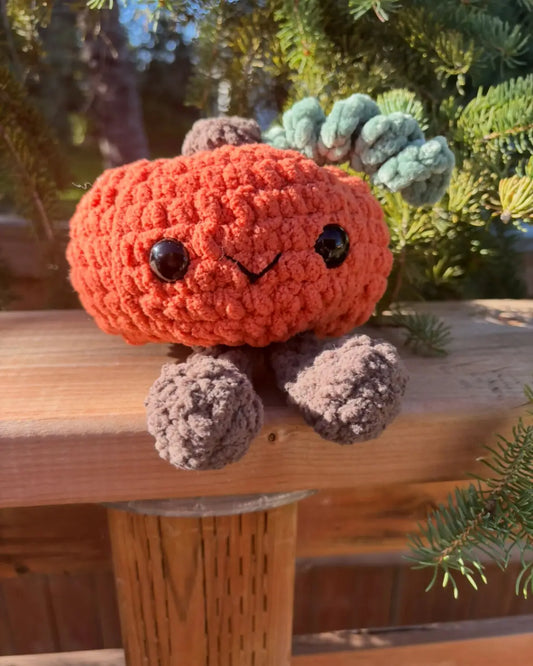 A cozy orange handmade crocheted pumpkin with legs featuring black button eyes and textured green arms, ideal for natural skincare canada gift displays