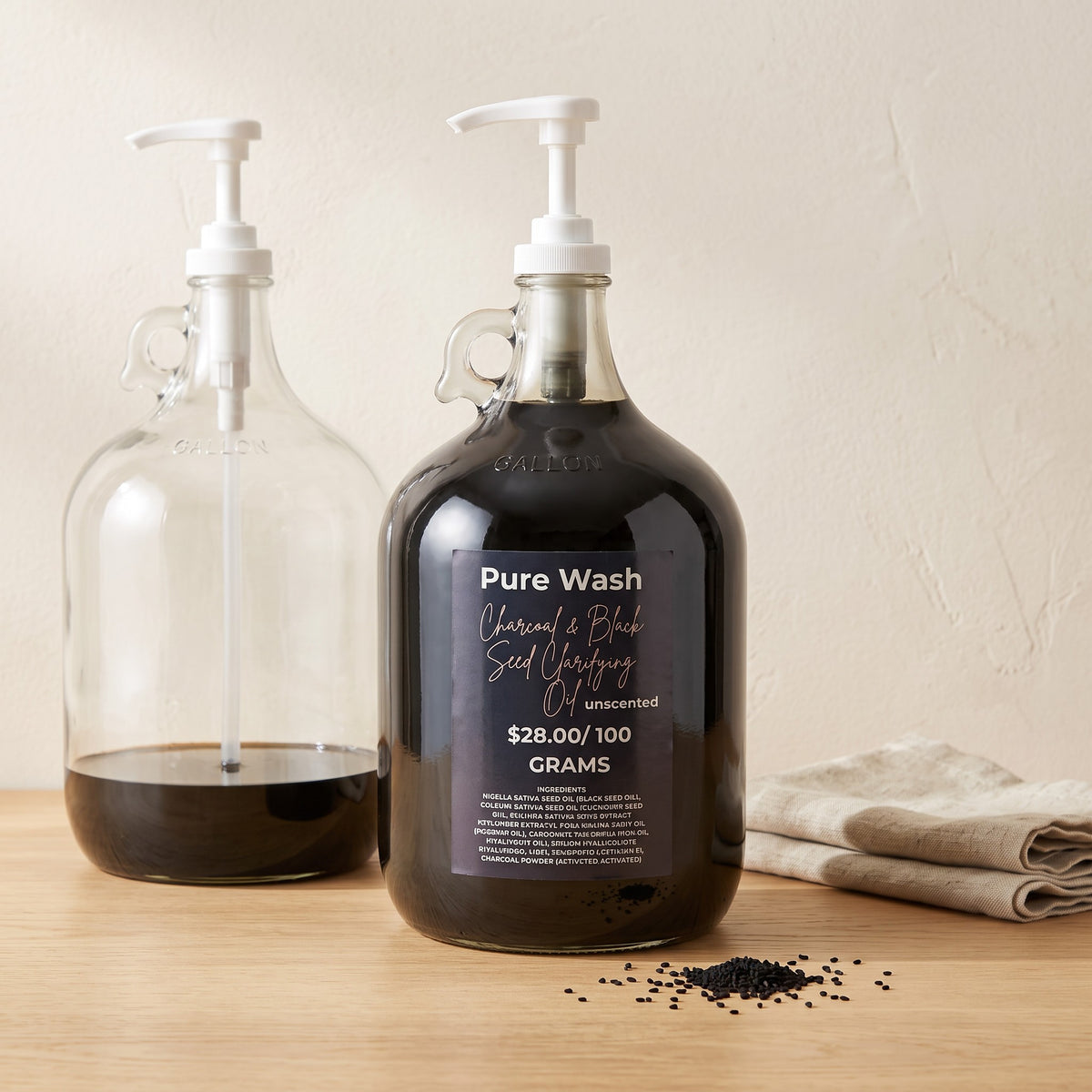 Pure Wash charcoal and black seed clarifying oil in a large glass refill jug with a pump dispenser on a wooden table.