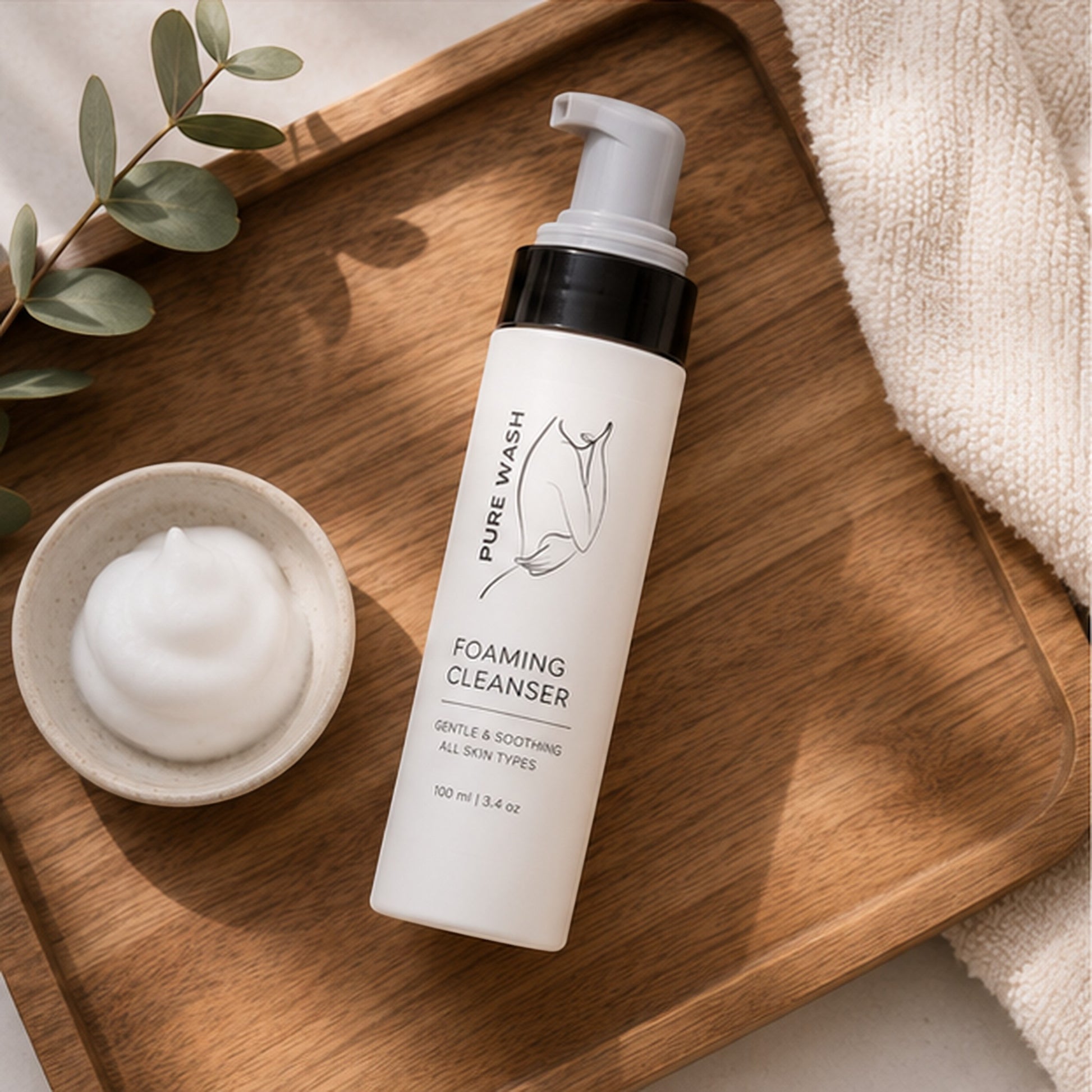 Pure Wash foaming hand soap bottle with a dollop of rich foam on a wooden tray with eucalyptus and a towel.