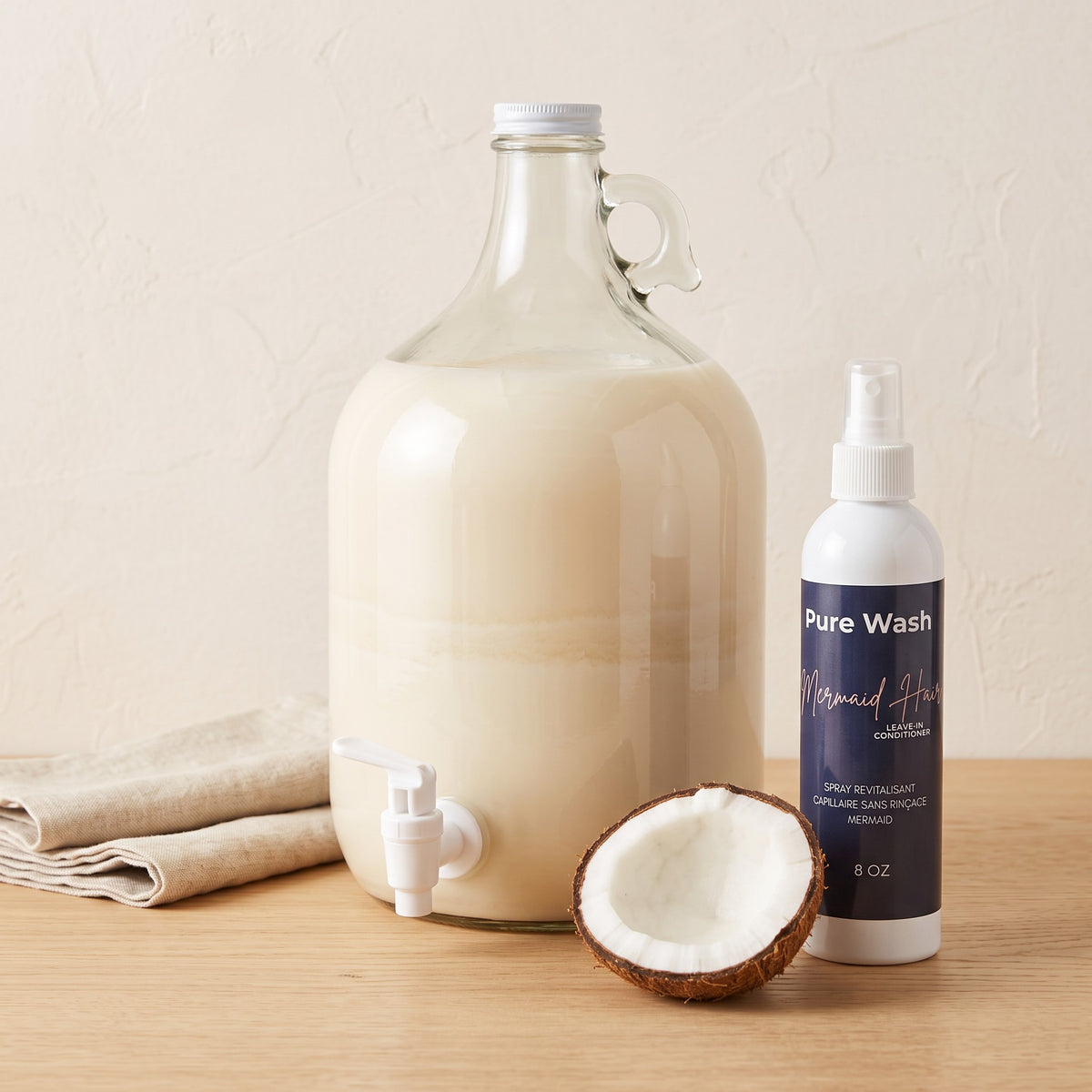 Pure Wash Mermaid Hair leave-in conditioner spray bottle next to a large glass dispenser and a fresh coconut half.
