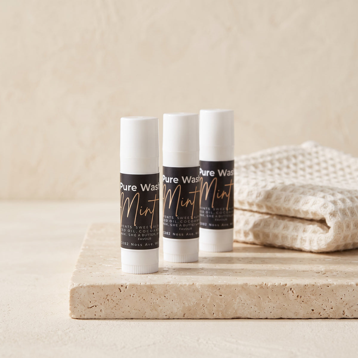 Three Pure Wash mint lip balm tubes on a stone surface next to a folded white waffle-knit towel.