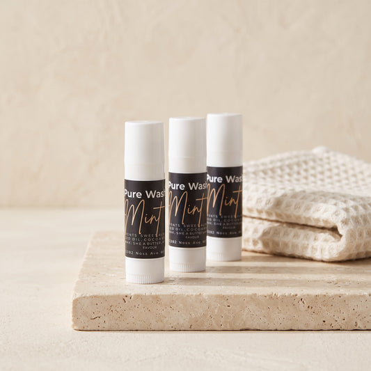 Three Pure Wash mint lip balm tubes on a stone surface next to a folded white waffle-knit towel.