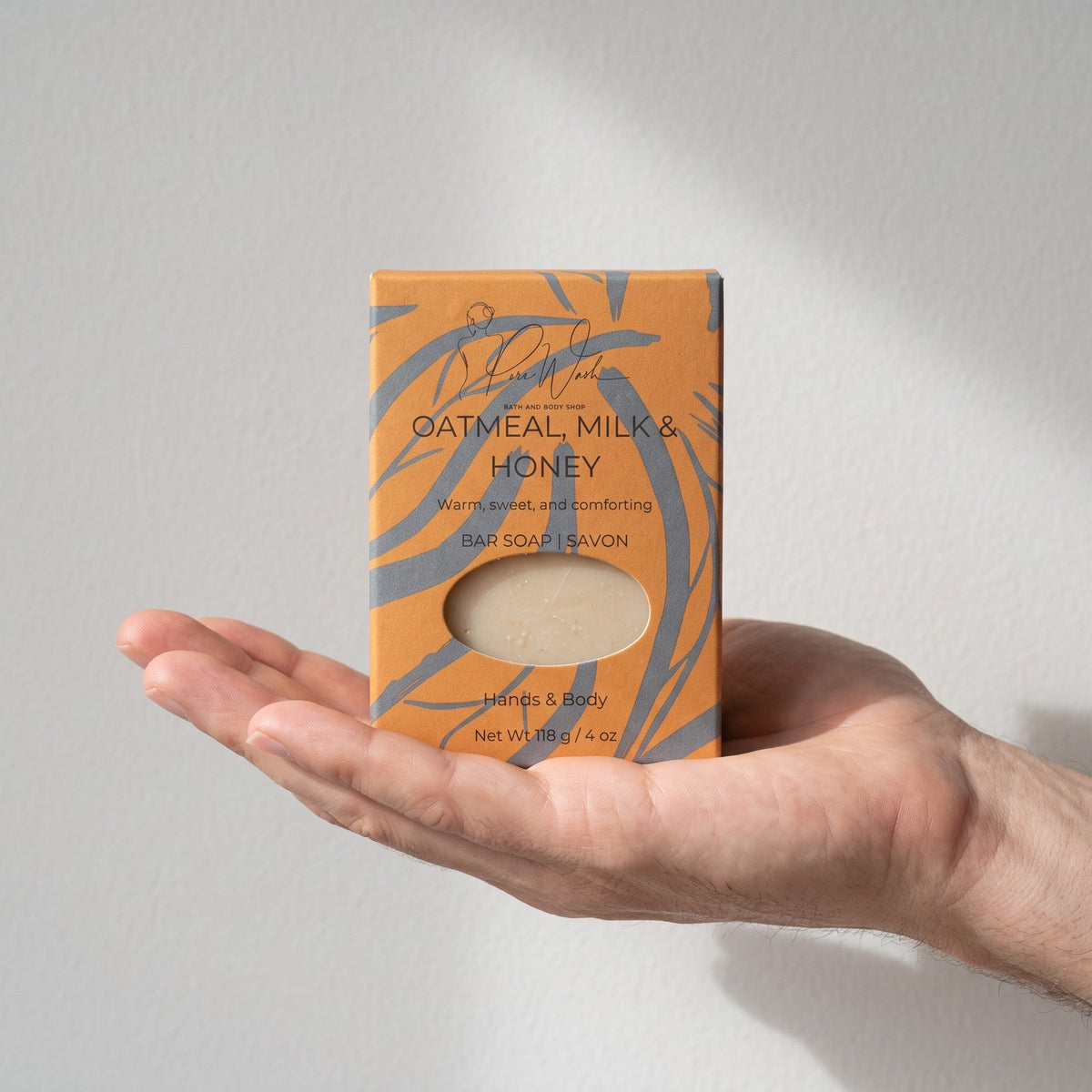 Pure Wash Oatmeal Milk & Honey soap bar in orange patterned packaging held in a hand against a neutral background.