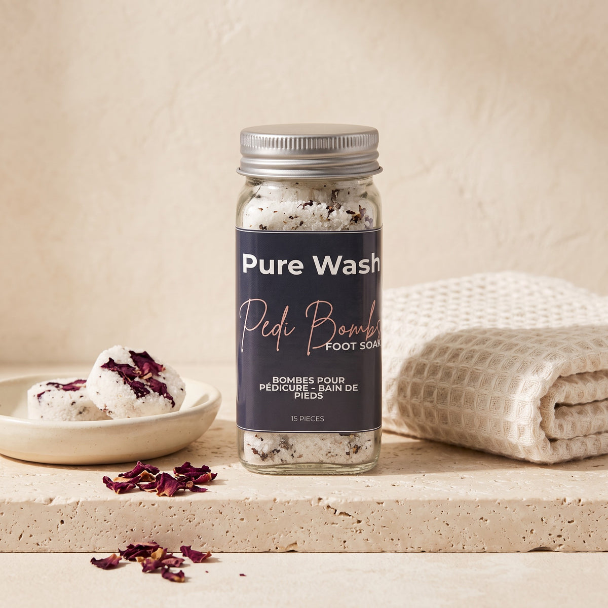 Pure Wash Pedi Bombs in a glass jar with dried rose petals on a stone surface next to a white towel