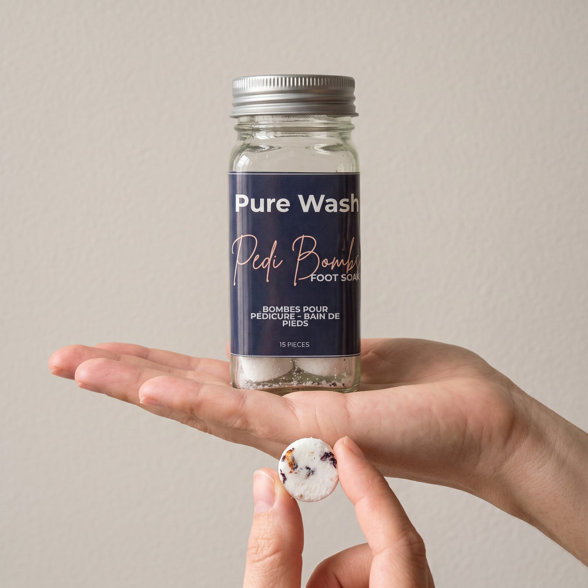 Pure Wash Pedi Bombs jar held in hand with a single floral-infused fizzing foot soak tablet in the foreground
