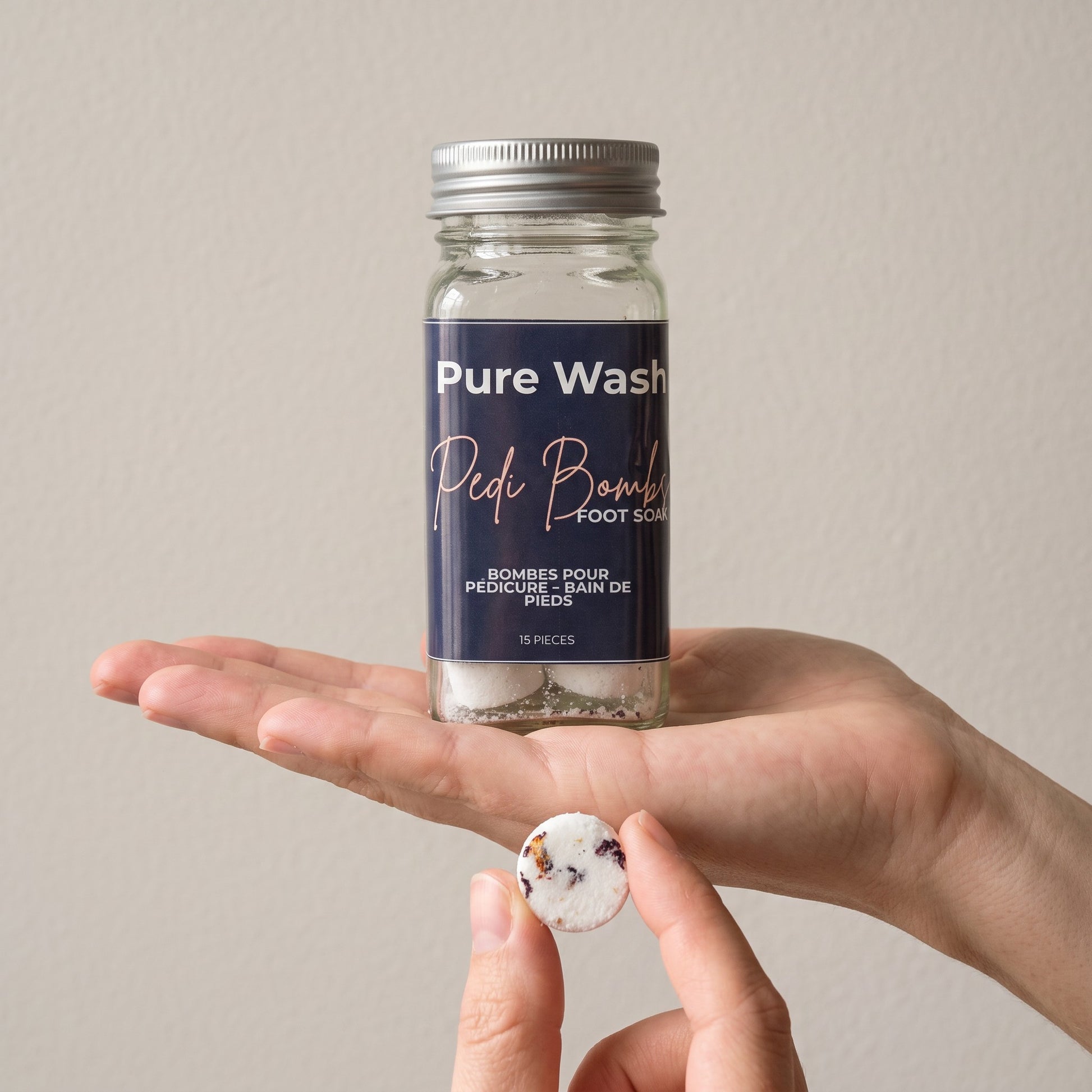 Pure Wash Pedi Bombs jar held in hand with a single floral-infused fizzing foot soak tablet in the foreground