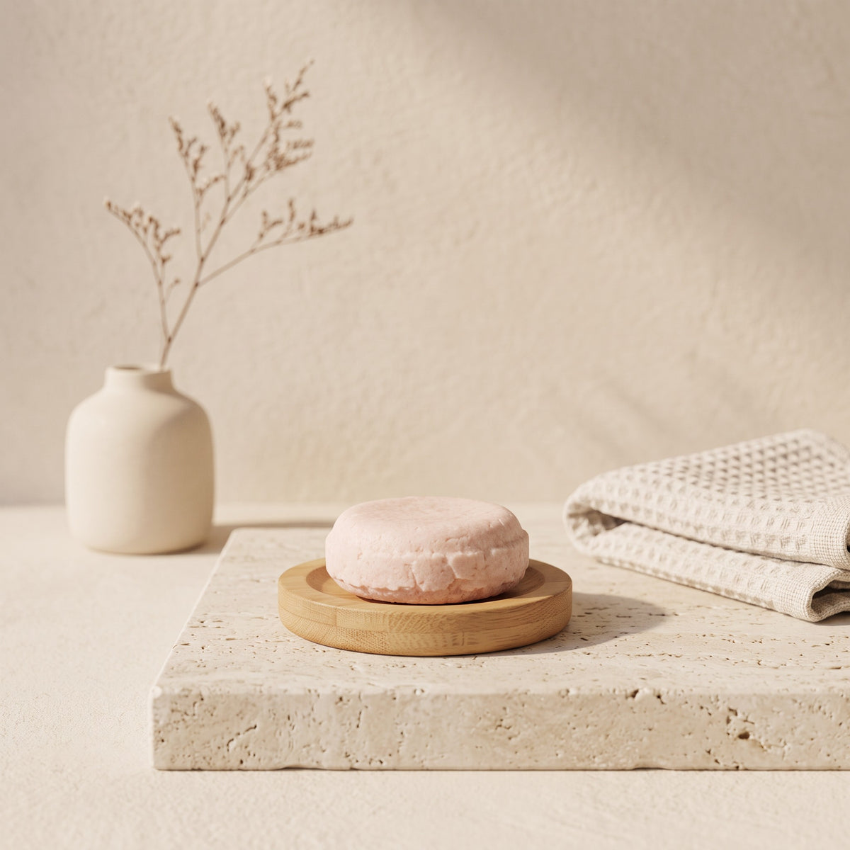 Pure Wash solid shampoo bar in soft pink on a wooden dish, set on a stone surface with a vase and waffle towel.