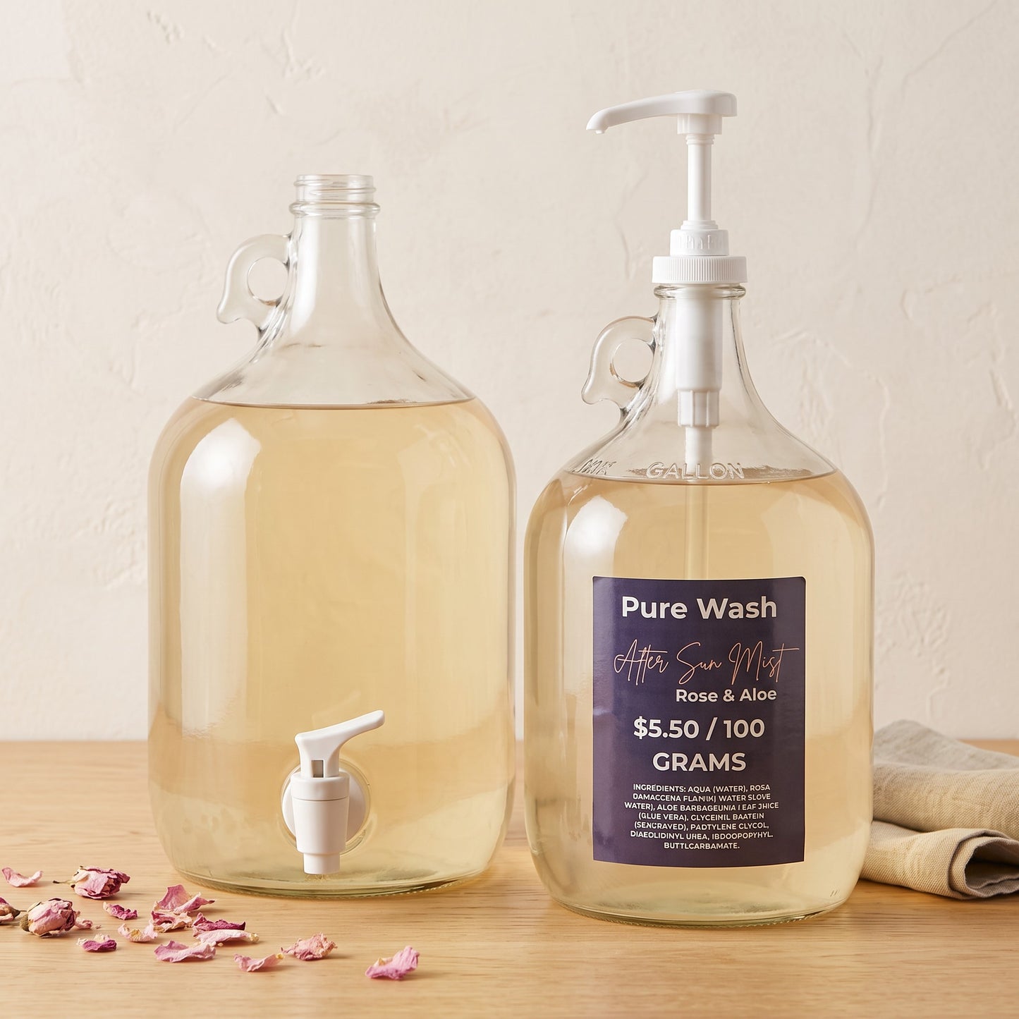 Pure Wash Rose & Aloe after-sun hydrating mist in large glass refill jugs with white pump and spigot on wood table.