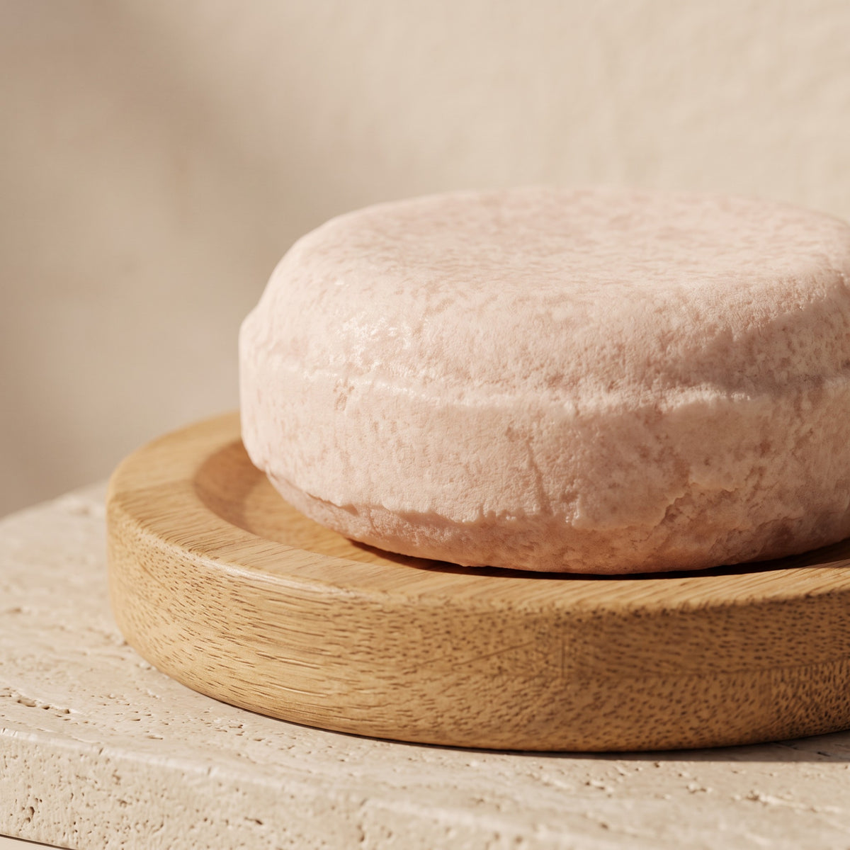 Pure Wash solid shampoo bar in a soft pink hue resting on a round wooden soap dish against a textured beige background