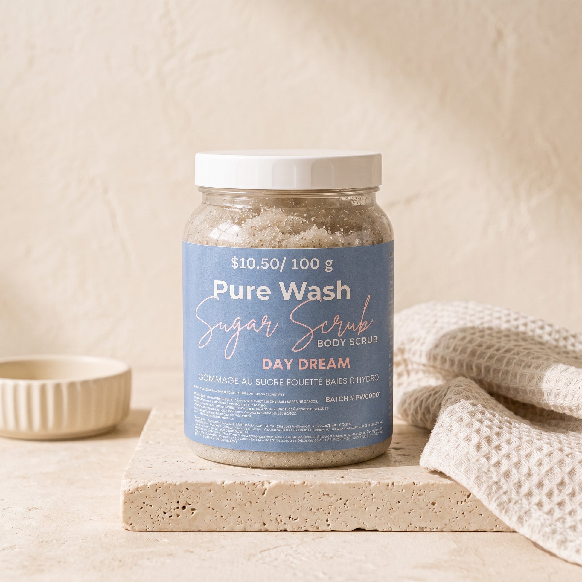 Pure Wash Day Dream sugar body scrub in a clear jar with a blue label, sitting on a stone pedestal with a towel.