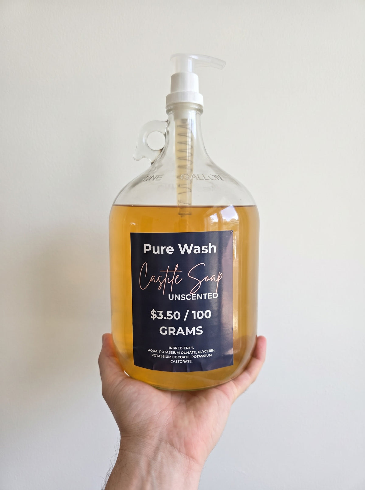 Pure Wash unscented concentrated liquid castile soap in a one-gallon glass jug held by a hand.