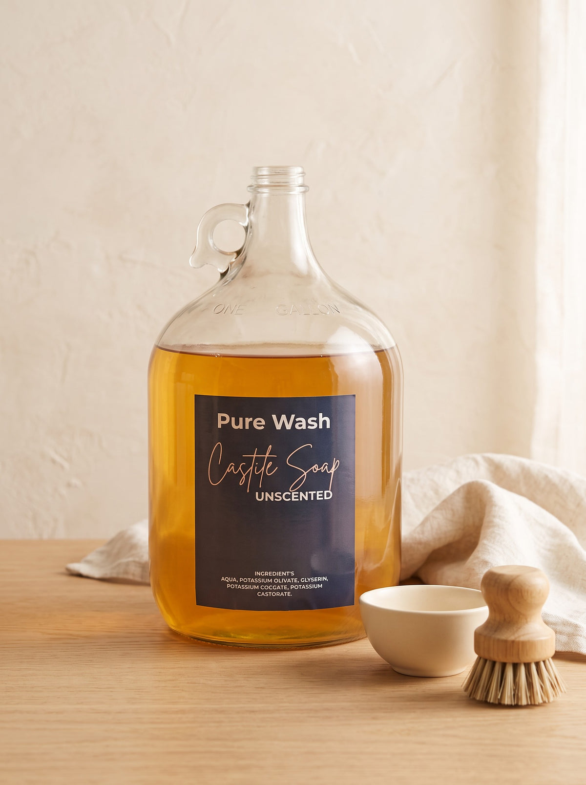 Pure Wash unscented concentrated castile soap in a one-gallon glass jug with a wooden dish brush on a wooden table.