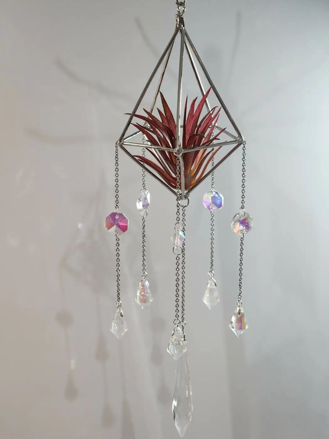 A geometric silver metal pyramid air plant suncatcher with a red air plant and iridescent crystal beads hanging
