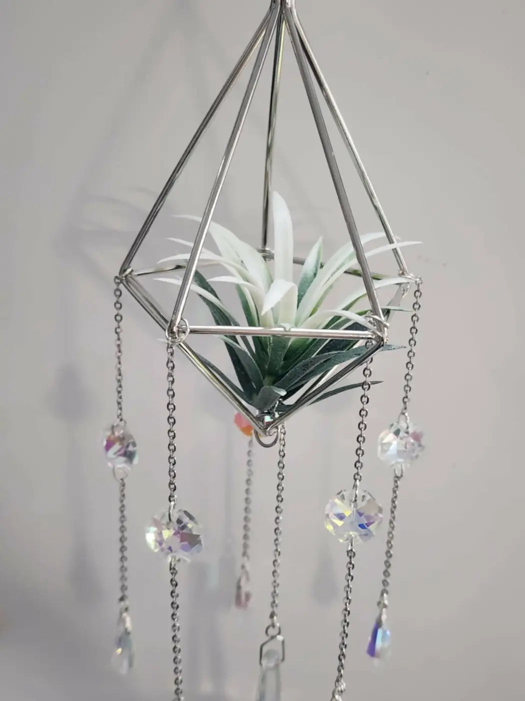 A geometric silver metal pyramid air hanging planter with dangling crystal chains holding a small air plant suncatcher