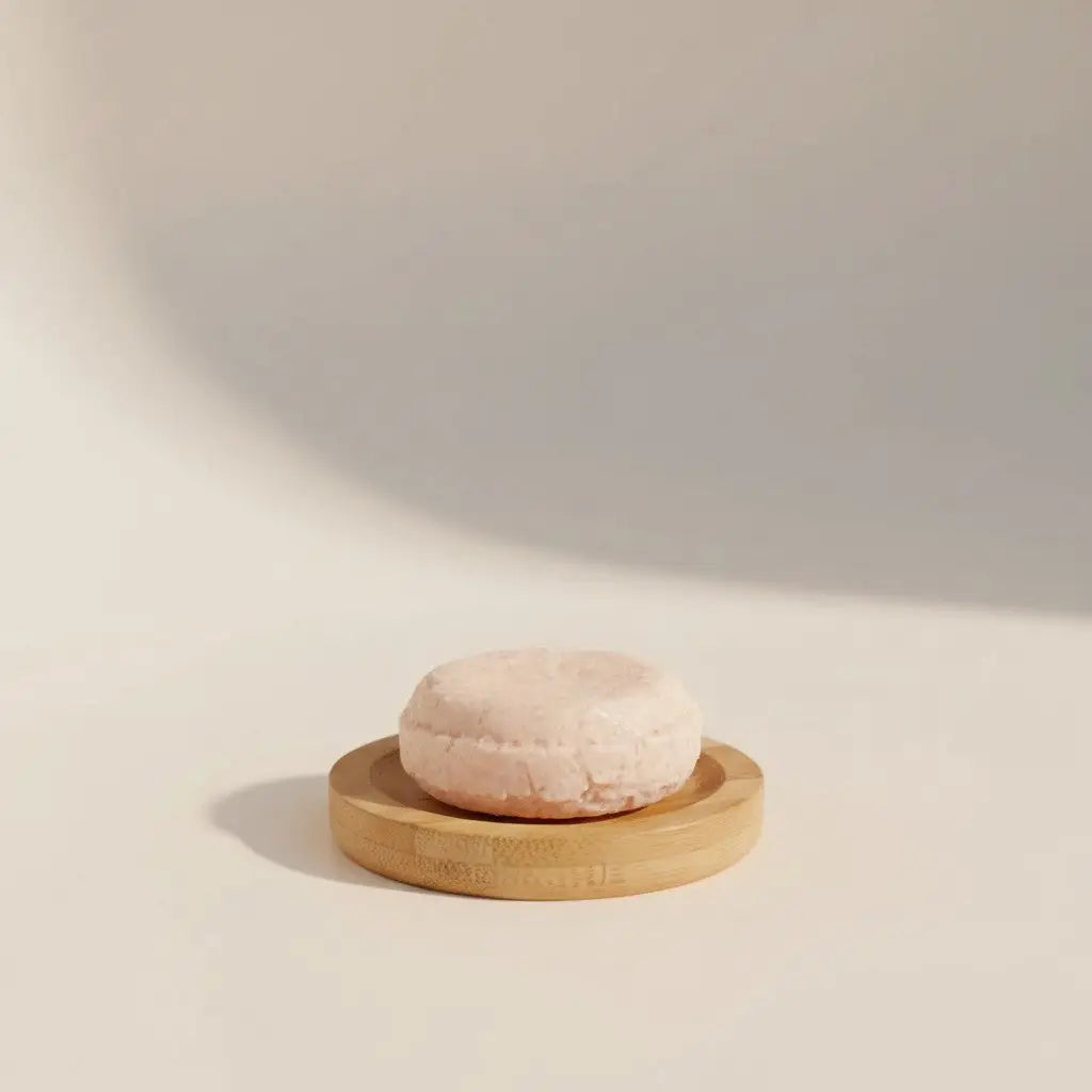 Pale pink Pure Wash shampoo bar with a textured surface on a light wood dish, for soft, easy-to-comb hair