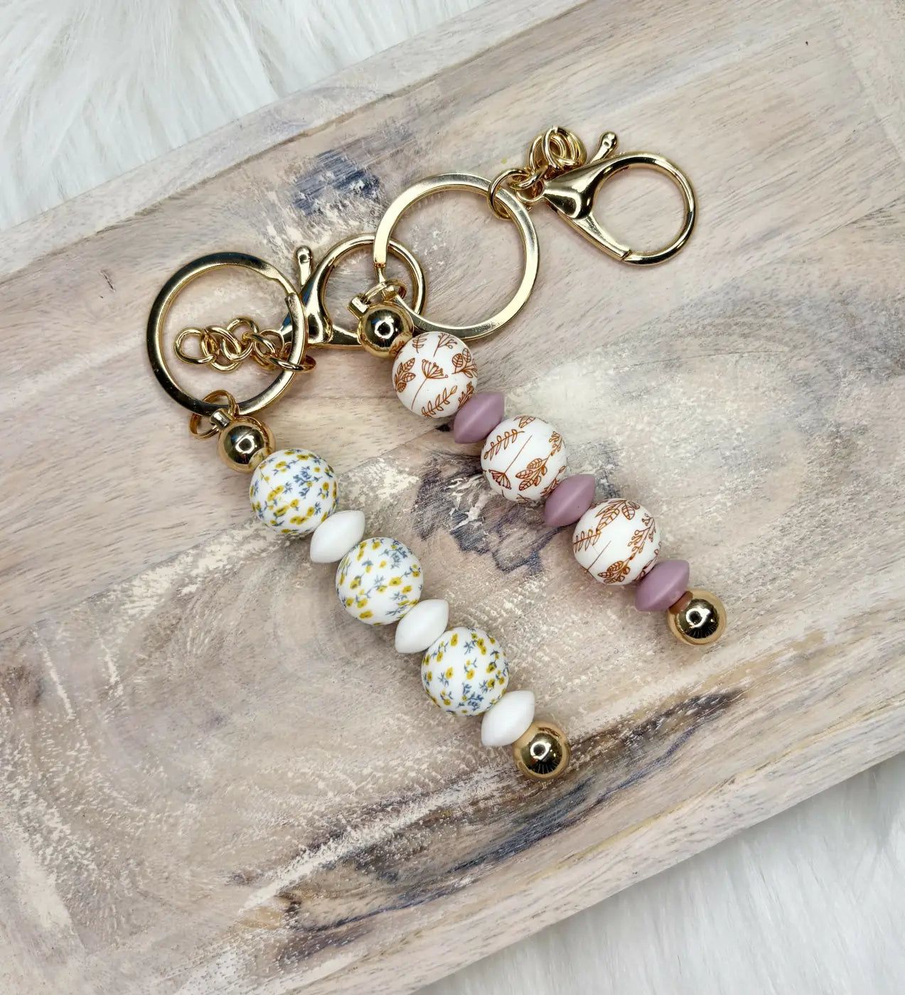 Two silicone bead keychains with gold clasps featuring white yellow floral and matte lavender beads for diaper bag essentials