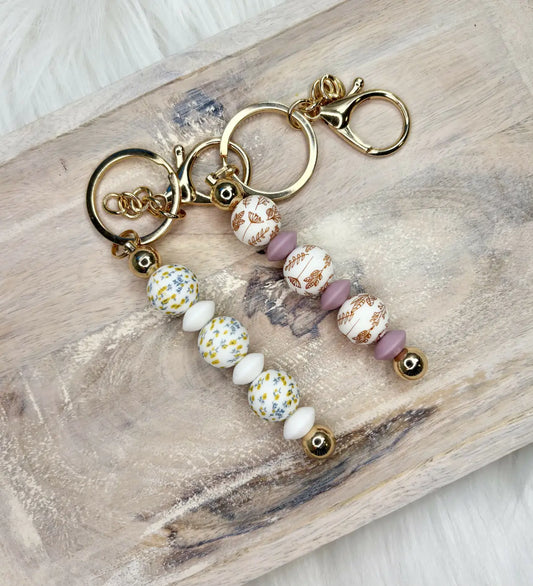 Two silicone bead keychains with gold clasps featuring white yellow floral and matte lavender beads for diaper bag essentials
