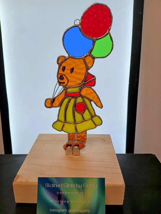 A handmade locally orange teddy bear stained glass ornament w balloons in yellow-green dress holding red blue green balloons