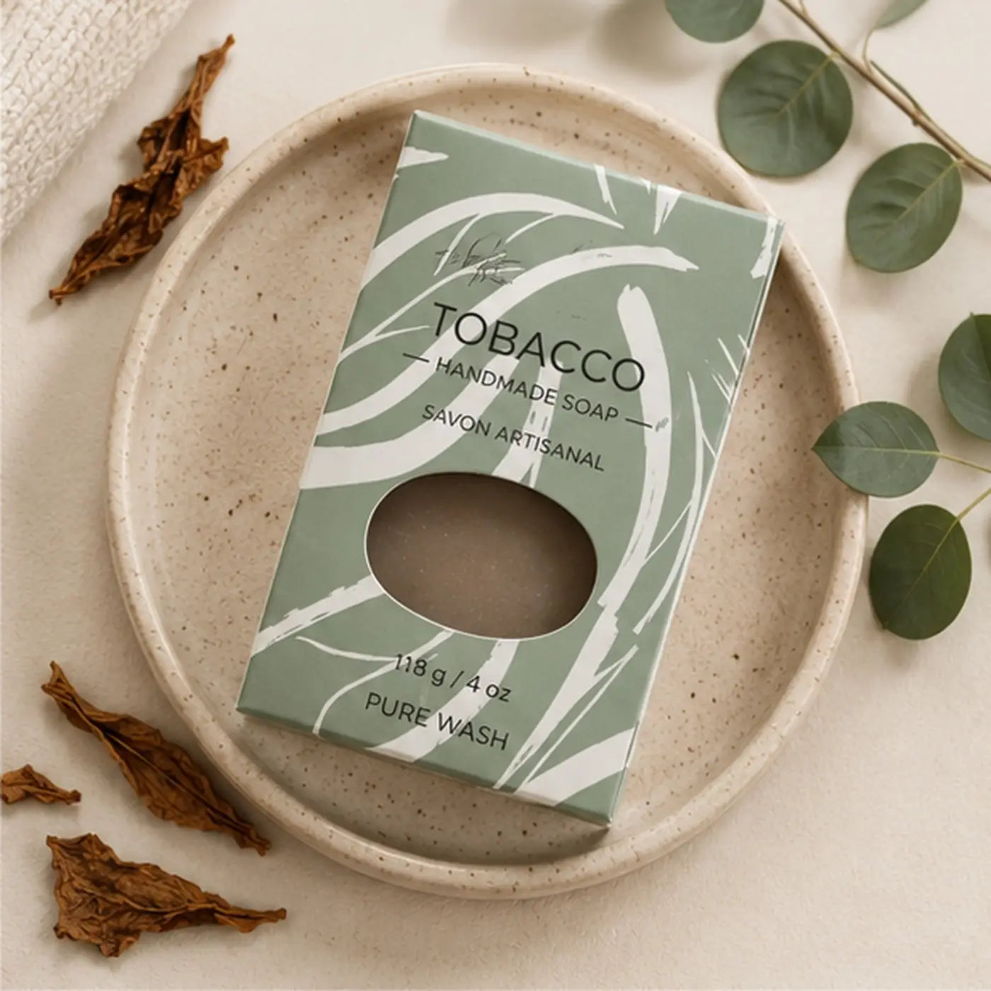 A matte sage green rectangular box with white leaf patterns and a clear window revealing handcrafted tobacco leaf soap without palm oil