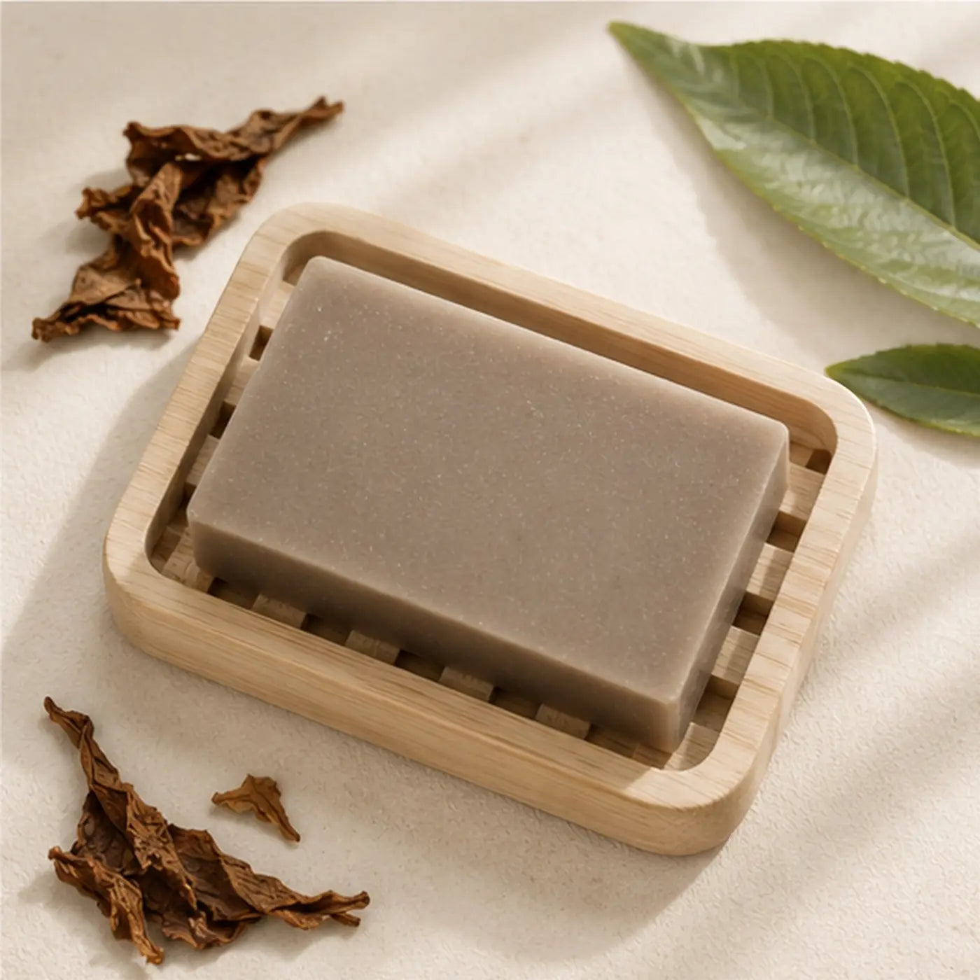 A rectangular matte gray tobacco leaf soap bar made with palm oil rests in a light wood-grain soap dish handcrafted skincare